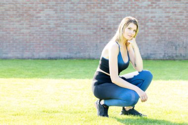 Uzun sarı saçlı, spor siyah ve mavi bluzlu ve taytlı formda genç bir kız çimenlerin üzerinde oturuyor ve gülümsüyor. Yüksek kalite fotoğraf