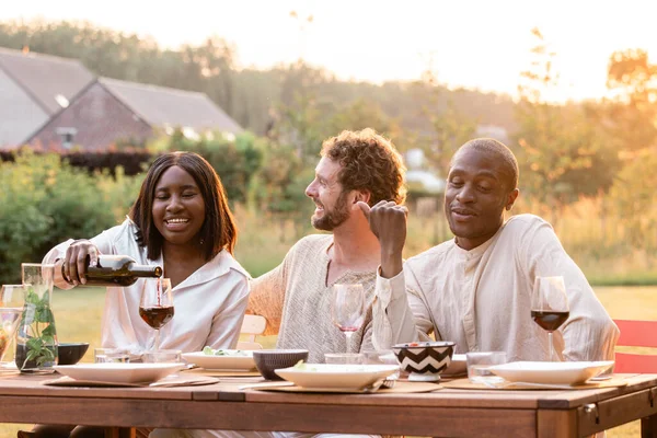 In the warm glow of an evening garden party, a diverse group of millennial friends come together around a beautifully set dinner table. They pour wine, clink glasses, and share in the joy of each