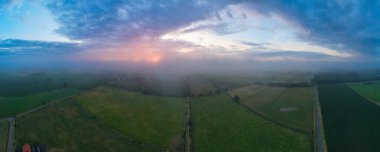 Güneşin kırsal bir arazide yükseldiği sisli ve bulutlu bir sabahın güzelliğini yansıtan hayranlık uyandıran bir hava manzarası. Her biri farklı ekinlere sahip çeşitli tarlalar, göz alabildiğince uzanıyor.