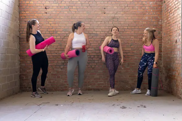 In the vibrant atmosphere of an industrial gym with a striking brick wall backdrop, a group of young and attractive sporty women, each with unique body shapes, come together. They share camaraderie