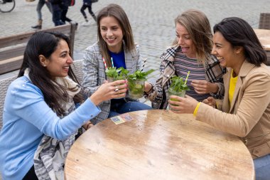 Kalabalık bir Avrupa şehir merkezinde bulunan bir kafede Mojito kokteylinin tadını çıkaran çok ırklı bir kadın arkadaş grubunun canlandırıcı bir görüntüsü. Sahne, dostluğu özetliyor.