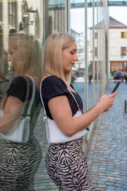 Beyaz çantalı, modaya uygun giyinmiş neşeli sarışın bir kadın şehirdeki bir ofis binasının cam duvarına yaslanıyor. Akıllı telefonuna dalmış, kayıyor ve büyük bir ilgiyle bakıyor.