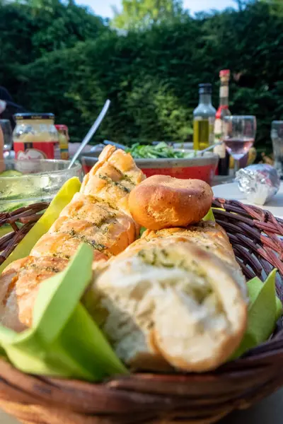 A rustic bread basket takes center stage on an outdoor garden table set for a barbecue. In the background, a delightful assortment of sauces, oil, drinks, and glasses of wine awaits. Bread Basket on
