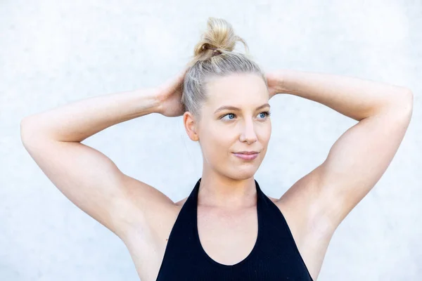 Capture the joy of relaxation and stretching with this footage. A young blonde woman stands against a serene concrete background, arms and hands gently behind her head and neck. With a content smile