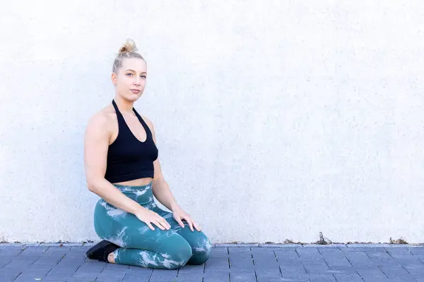 A young blonde Caucasian woman in black and blue sports apparel sits on her knees against a concrete wall, showcasing her sporty and active lifestyle with determination. Sporty Millennial Woman in