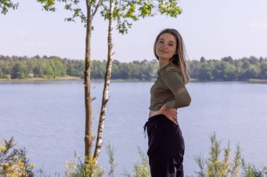 Güzel ve büyüleyici genç bir bin yıllık esmer, bir orman gölünün sakin kıyılarında duruyor. Yumuşak, doğal ışıkta yıkanmış, kameraya bakıyor, gözleri huzuru yansıtıyor.