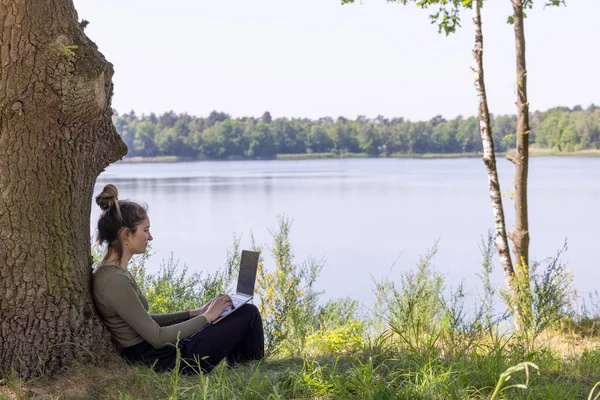 Embrace the freedom of remote freelance work with this captivating scene. A young woman, a dedicated freelancer, effortlessly balances her work and natures allure. She sits by a tranquil forest lake