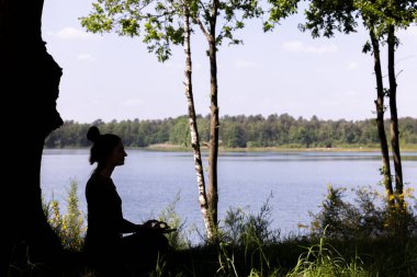 Genç bir milenyum kadınının dingin bir silueti yoga yapıyor ve sakin bir orman gölünün yanındaki yemyeşil çimenler üzerinde meditasyon yapıyor, doğayı kucaklıyor. Forest Lake 'de Yoga Meditasyonu. Yüksek