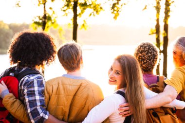 A heartwarming moment captured as a diverse group of five young friends embraces while enjoying a stunning sunset over a forest lake, bathed in golden light, with one smiling Asian friend looking back clipart