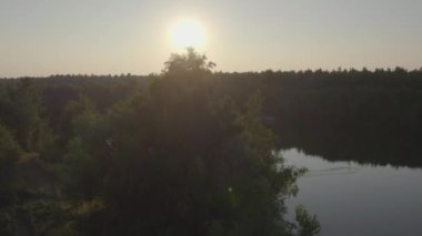 Kibar bir tırmanışta, tenha bir ağacın arkasındaki havadan bakıldığında sakin bir orman gölünün üzerinde nefes kesen bir günbatımını gözler önüne seriyor. Akşam gökyüzü kıpkırmızı renklerle boyandı.