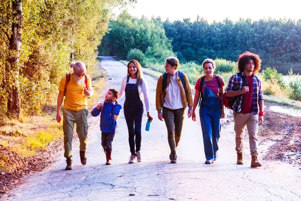 As the sun sets, casting a warm golden glow, a heartwarming scene unfolds on a quiet road by a forest lake. Friends of all ages, genders, and backgrounds come together, walking in tandem, their