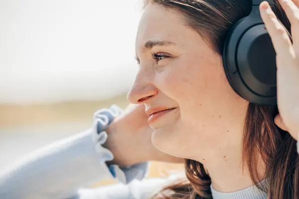 With the sea as her backdrop, a young woman in casual summer attire exudes excitement, her headphones a gateway to an auditory escape. The sunlight plays upon her features, highlighting a carefree