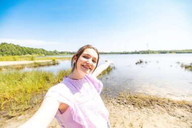 Büyük açık havada, mutlu genç bir kadın gölün kenarında selfie çeker, canlı gülüşü güneşin öptüğü bir günün özünü yakalar. Bu fotoğraf bir sağlık kutlamasıdır.