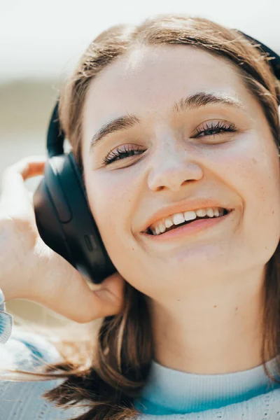 With the sea as her backdrop, a young woman in casual summer attire exudes excitement, her headphones a gateway to an auditory escape. The sunlight plays upon her features, highlighting a carefree