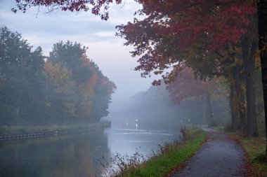 Görüntü, bir sonbahar sabahının yumuşak sisiyle kaplanmış sakin bir kanal görüntüsü yakalıyor. Su yolu canlı sonbahar renkleriyle bezenmiş ağaçlarla çevrili, kırmızı ve turuncu yaprakları ise...