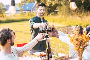 Bu görüntüde, bir grup arkadaş açık havada neşeli bir konuşmanın ortasında yakalanıyor. Bir adam ayakta şarap dökerken, diğerleri kadehlerini kaldırıp kutlama yaparken, onun hareketi dinamiktir.