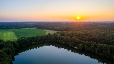 Bu görüntü, son ışığın tenha bir gölü süslediği alacakaranlığın huzurlu bir tasviri. Güneş, ateşli bir küre, ufukta süzülür, gökyüzünde sıcak renkler oluşturur. Çevreleyen
