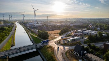 Görüntü, şehir manzarasını kesen bir kanalın havadan geniş bir görüntüsünü sunuyor, sabah güneşi sahnenin üzerine yumuşak, altın bir ışık saçıyor. Rüzgâr türbinleri ön planda duruyor