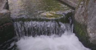 Görüntüler küçük ama dinamik bir şehir şelalesini yakalıyor. Burada su beton bir kanalda bir basamaktan aşağı yuvarlanıyor. Su hareketi sürekli akan bir enerji dansıdır, köpüklü bir köpük yaratır.