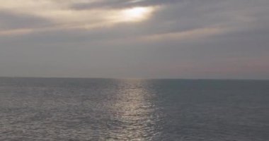 Durgun görüntüler, okyanusun engin ve dingin genişliğini bastırılmış bir gökyüzünün altında yakalıyor. Güneş ışınları bulutların arasından kırılıyor, su yüzeyinde parıldayan bir yol açıyor,