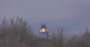 Bu sakin görüntüde, akşamları deniz feneri ışığının kıyı kum tepelerinin dalgalanan otları arasında ritmik bir şekilde dönmesiyle noktalanır. Yumuşak alacakaranlık renkleri doğayla harmanlanır.