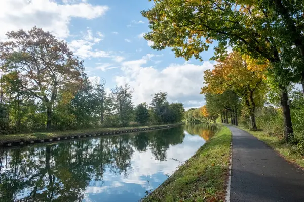 This tranquil image captures the essence of an autumn day beside a calm canal. A paved pathway guides the viewers eye through a scene framed by trees with leaves in shades of green and gold. The