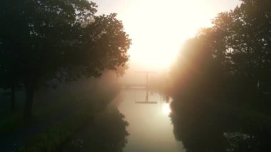 Bu anımsatıcı yükselen görüntü, yoğun bir sabah sisinin içinden süzülen güneşin sıcak parıltısını mistik bir atmosfer yaratarak gösteriyor. Ağaçların siluetleri çerçeveler güneş ışınları, döküm gölgeler