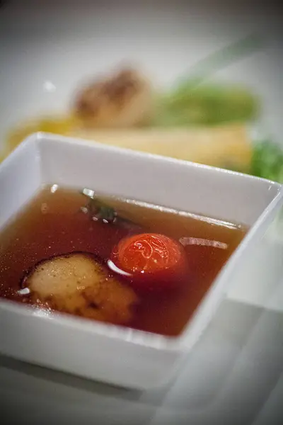 This image presents a deconstructed soup appetizer in a minimalist white bowl, highlighting the clarity of the broth and the precision of its components. A perfectly round tomato and a delicate slice