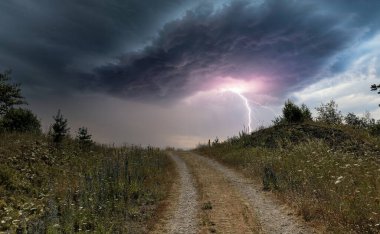 Bu görüntü, fırtınalı bir gökyüzünün kalbine uzanan tek bir toprak yolun olduğu kırsal bir ortamda dramatik bir fırtına yakalar. Yıldırımın mor ve pembe renkleri kara kara kara düşüncelere dalıyor.