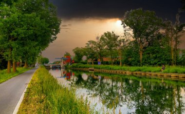 Bu sakin ama uğursuz görüntüde sakin bir kanal, yaklaşan fırtına bulutlarını ve akşam ışığının sonuncusunu yansıtır. Yol, su yoluna paralel gidiyor. Kırmızı, tuhaf bir çatıya doğru gidiyor.