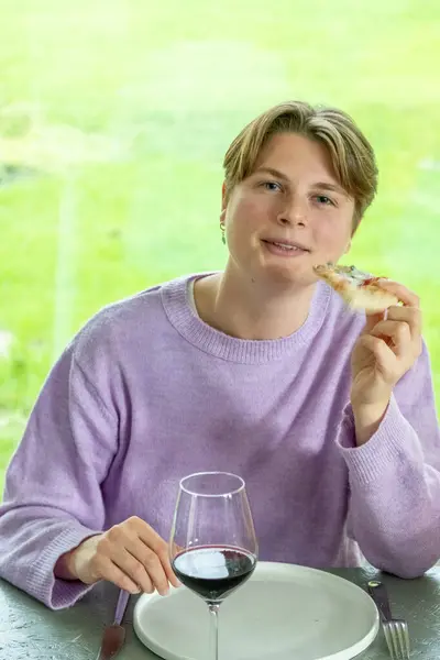 The photograph shows a person in a lilac sweater enjoying a meal, a slice of pizza in hand with a half-filled glass of red wine on the table. The individuals relaxed posture and contented expression