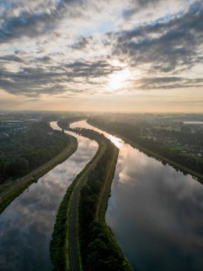 Bu hava fotoğrafı gündoğumunda bir nehrin barışçıl özünü yakalar. Su, yumuşak renklerin gökyüzünü yansıtırken yumuşak bir sis yüzeyin üzerinde süzülür ve sınırları harmanlar.