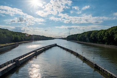 Bu geniş manzara, bulutlarla ve ışıl ışıl güneşle benekli bir gökyüzünün altındaki yemyeşil bir araziyi kesen bir kanalı yakalar. Kilit yapıları insan mühendisliğinin hünerlerini sergiliyor.
