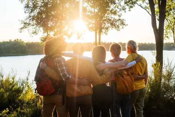 This heartwarming image showcases a group of friends, arms around each other, as they gaze at a lake bathed in the golden light of the setting sun. Their silhouettes against the radiant backdrop speak