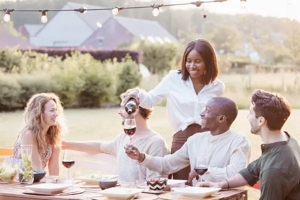 A Black woman stands pouring wine with a smile, as her friends, a Caucasian woman and two men, one Black and one Caucasian, look on with glasses in hand. Theyre seated around a rustic outdoor table