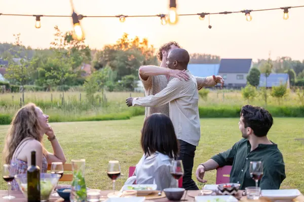 A heartwarming scene unfolds in the golden light of a setting sun, where two men, one Black and one Caucasian, share a hearty embrace, celebrating amidst a backyard dinner. Their friends, including a