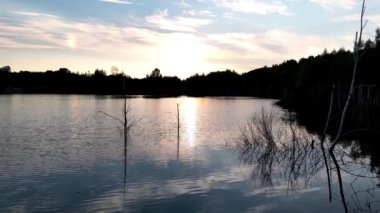 Üstgeçit görüntüsü, alacakaranlıktaki gölün sessiz güzelliğini yakalıyor. Su sakin, batan güneşin batan ışığını yansıtıyor. Sudan çıkan seyrek bitkilerin siluetleri ekliyor