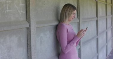 Günlük kıyafet giymiş genç bir kadın, şehir ortamında beton bir duvara karşı duruyor, akıllı telefonuyla mesajlaşıyor. Doğal ışık etrafında yumuşak gölgeler oluşturur.