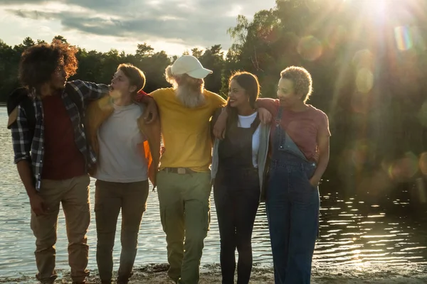 As the sun begins to set, casting a beautiful glow over the lake, a diverse group of friends share a serene moment. The group includes a Middle-Eastern man, a young Caucasian man, an elderly Caucasian