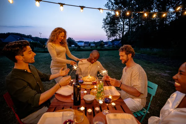 As dusk settles, an elegant outdoor dinner party is in full swing. A Caucasian woman stands to light a sparkler, adding magic to the moment, while a Black man seated at the table shares a laugh with
