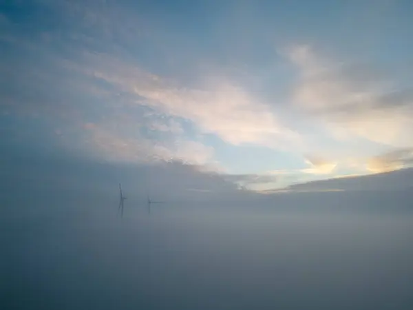 Captured with a drone above the mist, this image is a serene depiction of wind turbines emerging like giants from a sea of fog at dawn. The sky, a canvas of soft blues and warm pastel hues, suggests