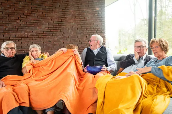 This heartwarming image captures a group of elderly friends enjoying each others company, comfortably seated on a sofa and covered with a bright orange blanket. The setting is a cozy room with a