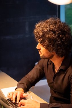 Resim, dikkatle dizüstü bilgisayarda çalışan odaklanmış bir bireyin profil görüntüsünü sunuyor. Kıvırcık saçları ve keskin kıyafetleri yazılım geliştirme işine dalmış yaratıcı bir profesyonele işaret ediyor.