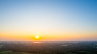 Bu büyüleyici görüntü, uçsuz bucaksız bir araziye sıcak bir parıltı saçan güneşin huzur verici güzelliğini gözler önüne seriyor. Ufuk turuncu ve mavi renklerin gradyanı ile süslenmiştir.