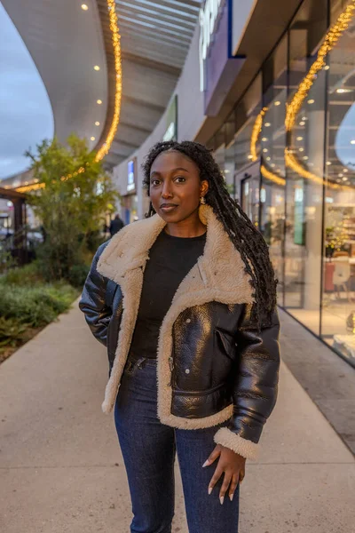 The photograph showcases a poised Black woman in an urban landscape during the evening. The setting sun casts a soft glow on the scene, illuminating her textured shearling coat and highlighting the