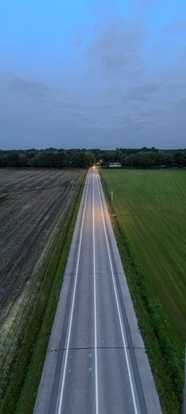 This vertical image showcases a country road taken at twilight, captured from an aerial view. The road creates a striking vanishing point, illuminated by the headlights of a lone vehicle in the