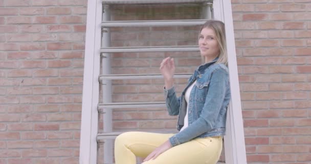 在这个慢镜头中 一个时髦的女人坐在城市的楼梯上 穿着斜纹棉布夹克和醒目的黄色裤子 散发着自信和时尚感 她轻松自在 — 图库视频影像