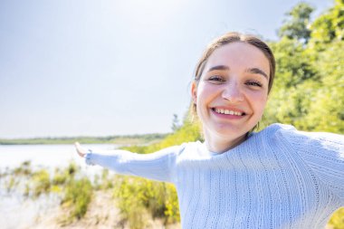 Görüntü, açık mavi süveter giyen neşeli genç bir kadını, sanki göl manzaralı bir yerde uzaktan bir şey gösteriyormuş gibi kolunu uzatarak yakalıyor. Parlak gülüşü ve açık jesti