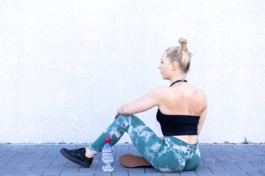 Bu görüntüde, yoga minderinin üzerinde beyaz bir duvara yaslanmış, antrenmanına ara vermiş genç bir kadın görülüyor. Siyah spor sutyen ve yeşil kamuflaj da dahil olmak üzere spor kıyafeti giyiyor.