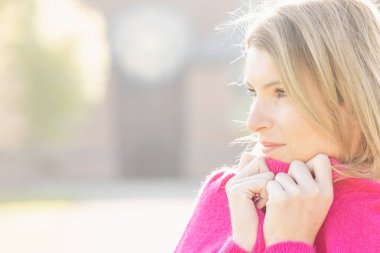 Bu görüntü, canlı pembe bir süveter giyen, düşüncelere dalmış genç beyaz bir kadını yakalıyor. Arka plandaki yumuşak odak sıcak ve sisli bir sonbahar gününün tipik ışıklarıyla sahneyi yıkıyor. Kadın.